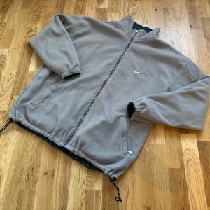 1990’s Vändbar fleece- och vindjacka från Nike ACG - Säljer en superpraktisk vändbar jacka från Nike! Ena sidan är en mysig grå fleece och andra sidan är en stilren mörkblå vindjacka. Perfekt för både höst och vår. Jackan har dragkedja och elastiska ärmslut. Den är i bra skick bortsett från liten liten defekt i ärmmudd som ses på bild.