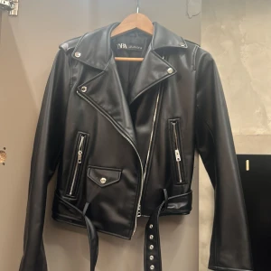 Svart skinnjacka från Zara - Säljer en snygg svart skinnjacka från Zara i storlek L. Jackan har en cool bikerstil med dragkedjor och bälte i midjan. Perfekt för höst och vår, och funkar lika bra till fest som till vardags. Den är i mycket bra skick och redo för nya äventyr! Ny skick, använd 2-3 gånger! 300 kr + frakt, nypris 600 kr