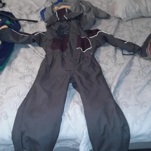 Grå overall med huva - Säljer en grå overall med huva, perfekt för kalla dagar. Den har vita detaljer och elastiska muddar vid ärmslut och benslut för extra skydd mot väder och vind. Overallen är i bra skick och har en praktisk dragkedja framtill.