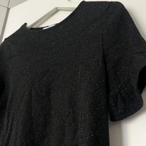 Svart glittrig topp från NA-KD - Svart glittrig topp från NA-KD i storlek XS. Korta ärmar med volangdetaljer. Älskar denna men tyvärr för liten för mig 🥹🪩