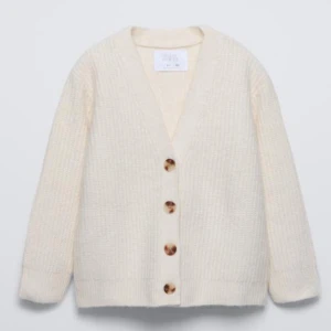 Vit ribbad kofta från Zara - Mysig vit kofta från Zara i ribbat material. Den har en v-ringad design med stora knappar framtill. Perfekt för kyliga dagar och kan enkelt matchas med olika outfits. Koftan har en avslappnad passform och långa ärmar.