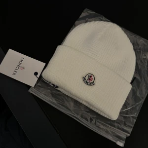 Vit stickad mössa från Moncler - Säljer en snygg vit stickad mössa från Moncler. Den är i nytt skick och har Monclers ikoniska logga broderad framtill. Perfekt för kalla vinterdagar och ger en stilren look. Mössan är mjuk och bekväm, vilket gör den till ett måste i garderoben. Kommer med originalförpackning och etikett.