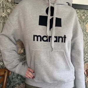 Grå hoodie från Isabel Marant - Säljer en supermjuk och bekväm grå hoodie från Isabel Marant. Den har en stor svart logga på framsidan och en praktisk magficka. Perfekt för kyliga dagar eller en avslappnad stil. Passar till allt och är ett måste i garderoben! 🖤
