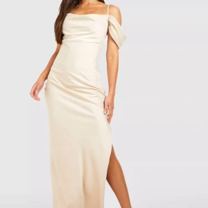Champagnefärgad långklänning - Elegant champagnefärgad långklänning i satin med offshoulder-design och smala axelband. Klänningen har en smickrande slits och en figurnära passform, perfekt för festliga tillfällen.