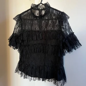 Svart spetsblus från By Malina - Säljer en elegant svart spetsblus från By Malina i storlek S. Blusen har korta ärmar och vackra volangdetaljer. Perfekt för festliga tillfällen eller en utekväll. Den är i mycket bra skick!