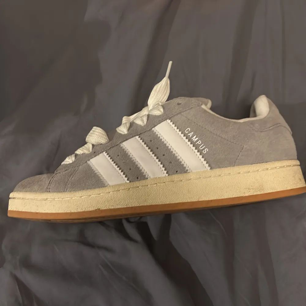 Säljer ett par snygga grå Adidas Campus sneakers i mocka. De har de klassiska tre vita ränderna på sidan och kommer med extra skosnören. Perfekta för en casual look och passar till nästan allt. Skorna är i mycket bra skick och levereras i originalkartong.. Skor.