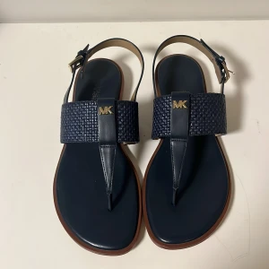 Blå sandaler från Michael Kors - Snygga blå sandaler från Michael Kors med flätad rem och guldfärgad MK-logga. De har en bekväm passform och justerbar rem runt ankeln. Perfekta för sommardagar och strandhäng. Använda men i bra skick.
