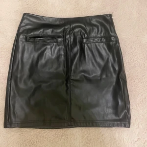 Svart skinnkjol - Snygg svart skinnkjol i kort modell. Perfekt för en kväll ute eller en cool vardagslook. Kjolen har en dragkedja på sidan och stilrena fickdetaljer. Passar perfekt till både höga klackar och sneakers.