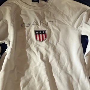 Vit långärmad tröja från GANT - Säljer en stilren vit långärmad tröja från GANT. Den har ett broderat märke på bröstet med en sköld i rött, vitt och blått. Perfekt för en casual look eller en avslappnad dag ute. Tröjan är i bra skick och redo för en ny ägare!