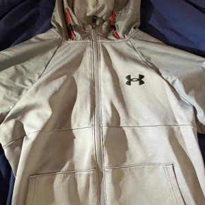 Grå hoodie från Under Armour - Säljer en grå hoodie från Under Armour i mycket bra skick. Den har en dragkedja framtill och en justerbar huva med röda detaljer. Perfekt för höst och vår, och passar både till träning och vardag. Superbekväm och stilren design!