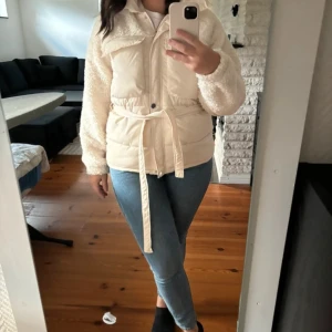 Beige dunjacka med bälte - Säljer en supermysig beige dunjacka med bälte i midjan. Jackan har en dragkedja framtill och är perfekt för höst och vinter. Den har en fluffig look med  detaljer och är både stilren och varm. Perfekt för att hålla sig varm och snygg under kyliga dagar! Jackan är i storlek S och är normal i storlek, köpt för 699kr