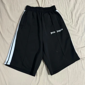 Svarta shorts från Palm Angels - Säljer ett par svarta shorts från Palm Angels i storlek M.  De är väldigt oversized. De har vita ränder längs sidorna och en elastisk midja för extra komfort. Perfekta för en avslappnad stil eller träning. Materialet är en blandning av bomull och elastan vilket ger en skön passform.