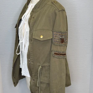 Olivgrön jacka med broderade detaljer - Säljer en olivgrön jacka med coola broderade detaljer på ärmen. Jackan är aldrig använd enbart en gång. Jackan har en dragkedja och knappar framtill, samt praktiska fickor med knappar och snörning i midjan. Perfekt för höst och vår!