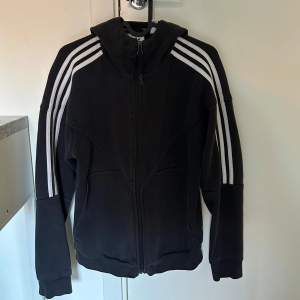 Säljer en svart huvtröja från Adidas i bra skick. Den har de klassiska tre vita ränderna längs ärmarna och en dragkedja framtill. På baksidan finns Adidas-loggan och text i vitt. Perfekt för en sportig look eller chill vardagsstil.