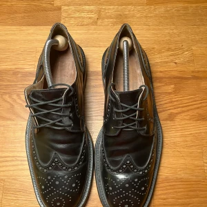 Svarta läderskor med broguemönster märke Lloyds - Snygga svarta läderskor i mycket bra skick med klassiskt broguemönster. Perfekta för både formella och semi-formella tillfällen. Skorna har snörning och en robust sula för extra komfort. Passar utmärkt till höst och vår.