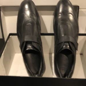 Svarta loafers från Brian Cress - Snygga svarta loafers i skinn från Brian Cress. Perfekta för både formella tillfällen och vardagsbruk. De har en stilren design med en diskret rem över vristen. Skorna är i mycket bra skick och kommer i originalkartong.