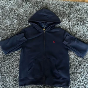 Mörkblå hoodie från Ralph Lauren - Säljer en snygg mörkblå hoodie från Ralph Lauren i storlek 14-16. Den har en dragkedja framtill och en klassisk röd logga på bröstet. Perfekt för en avslappnad stil och passar bra till både höst och vår. Huvan ger extra mysfaktor! 🧢Storleken passar ungefär som S, lite mindre 