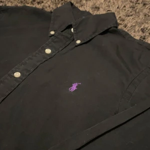 Svart skjorta från Ralph Lauren - Säljer en snygg svart skjorta från Ralph Lauren i storlek S samt slim fit. Skjortan är gjord av bomull och har en lila broderad logga på bröstet. Perfekt för både vardag och fest! 