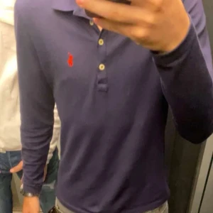 Mörkblå pikétröja från Ralph Lauren - Säljer en snygg mörkblå pikétröja från Ralph Lauren. Den har långa ärmar och en klassisk krage med knappar. Perfekt för både vardag och lite finare tillfällen. Den röda loggan på bröstet ger en stilren touch. Passar bra till jeans eller chinos!