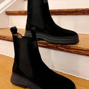 Svarta Chelsea boots i mocka - Snygga svarta Chelsea boots i mocka med en robust sula. Perfekta för höst och vinter, dessa boots har en klassisk design utan snörning, vilket gör dem lätta att ta på och av. De är i nytt skick och redo att ge din outfit en stilren touch.