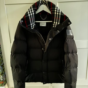 Svart dunjacka från Burberry - Säljer en snygg svart dunjacka från Burberry i mycket bra skick. Jackan har en klassisk rutig krage, dragkedja och knappar framtill, sidofickor med dragkedja samt en stilren logga på ärmen. Även två innerfickor med dragkedja. Varm!