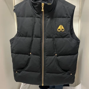 Svart dunväst från Moose Knuckles 24k pläterad badge - Säljer en svart dunväst från Moose Knuckles i mycket bra skick. Västen har en snygg guldfärgad dragkedja och ett coolt märke på bröstet. Sen finns det en del andra guld detaljer jag säkert har missat. Perfekt för höst och vinter, med en quiltad design som håller dig varm. Passar både till vardags och fest!