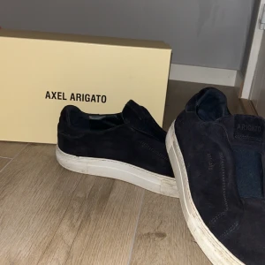 Mörkblå sneakers från Axel Arigato - Säljer ett par snygga mörkblå sneakers från Axel Arigato i mocka. De har en stilren design med vit sula och är i bra skick. Perfekta för både vardag och fest! Kommer med originalförpackning och dustbag. Skorna är lite smutsiga men det går enkelt att tvätta bort! Endast använda fåtal månader men använt sparsamt.
