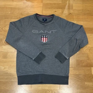 Grå sweatshirt från GANT - Säljer en snygg grå sweatshirt från GANT med ett emblem på bröstet. Tröjan har långa ärmar och ribbade muddar vid ärmslut och nederkant. Perfekt för en avslappnad stil eller en kylig dag. Passar både till vardags och lite mer uppklädda tillfällen.