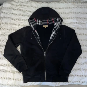 Svart kofta från Burberry - Säljer en snygg svart kofta från Burberry i storlek M.Kan gå ner i pris vid snabbaffär, Den har en klassisk rutig insida i huvan och dragkedja framtill. Perfekt för höst och vår, med långa ärmar och mjukt bomullsmaterial. Passar både till vardags och lite finare tillfällen. 🖤
