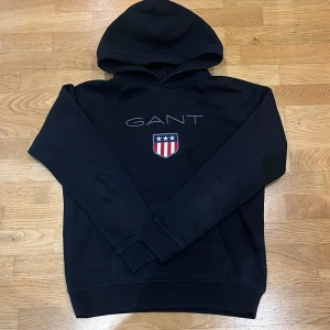 Svart hoodie från GANT - Säljer en snygg svart hoodie från GANT med deras ikoniska logga och ett sköldmärke på bröstet. Perfekt för både vardag och chill. Den är i mycket bra skick och superbekväm med en klassisk passform. Passar perfekt till jeans eller joggers!
