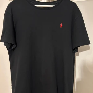 Svart t-shirt från Ralph Lauren - Säljer en klassisk svart t-shirt från Ralph Lauren i bomull. Den har en liten röd logga på bröstet och rund halsringning. Perfekt för en stilren och avslappnad look. Passar till både vardag och fest!
