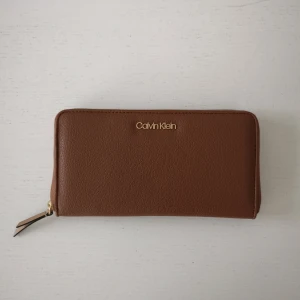 Brun plånbok från Calvin Klein - Säljer en snygg brun plånbok från Calvin Klein i mycket ny skick. Den är gjord av skinn och har en praktisk dragkedja runtom. Insidan har flera fack för kort och en mittficka med dragkedja för mynt. Perfekt för att hålla ordning på dina grejer med stil!