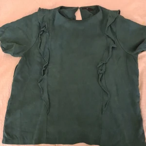 Grön blus med volangdetaljer - Säljer en fin grön blus med volangdetaljer längs framsidan. Blusen har korta ärmar och en liten öppning i ryggen. Perfekt för både vardag och festliga tillfällen. 💚