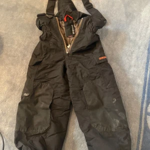 Svarta vinterbyxor från Didriksons - Säljer ett par svarta vinterbyxor från Didriksons i storlek 110. Byxorna har justerbara hängslen och dragkedja framtill. Perfekta för kalla vinterdagar med slitstarkt material och elastiska benslut för att hålla snön ute. Använda men i bra skick, redo för nya äventyr i snön! ❄️