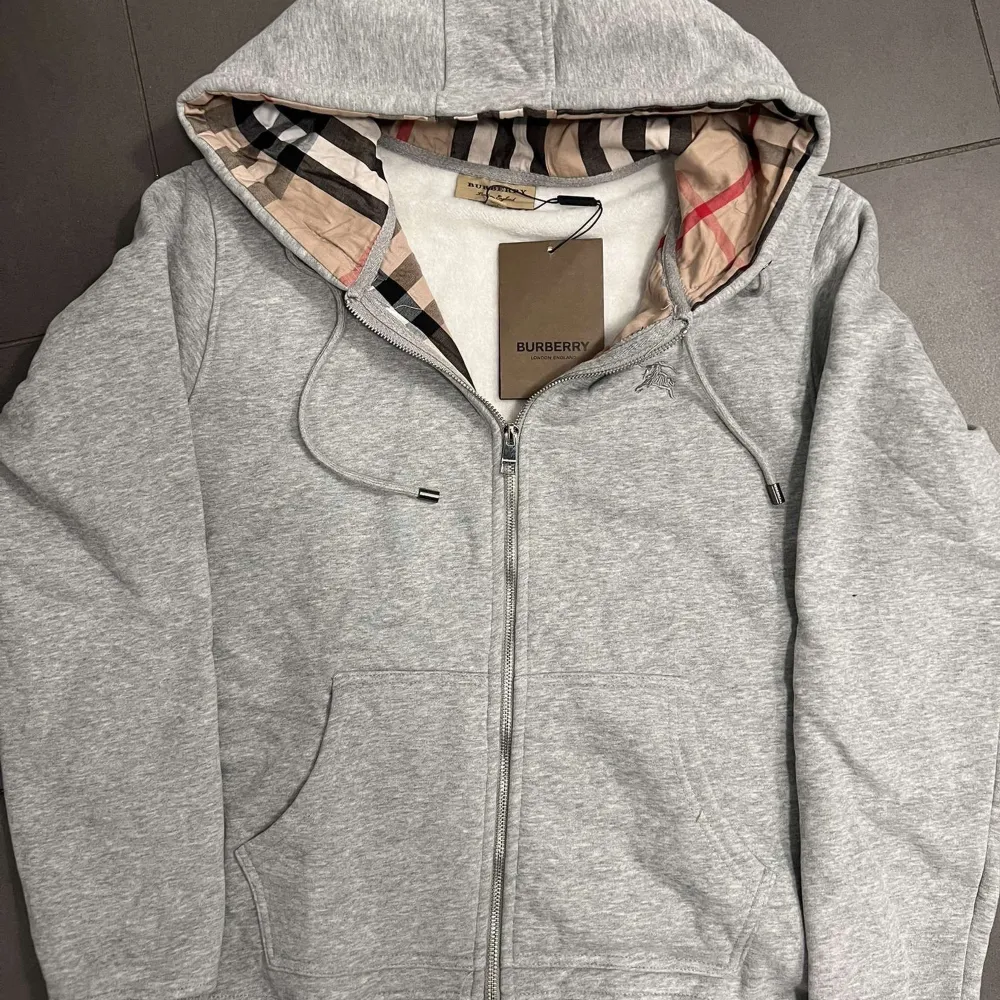 Säljer en snygg grå hoodie från Burberry i nytt skick. Den har en klassisk dragkedja och den ikoniska Burberry-rutiga fodringen i luvan. Perfekt för höst och vår, med en stilren broderad logga på bröstet. Passar både till vardags och lite finare tillfällen. Obs. Priset är ej hugget på sten❗️. Tröjor & Koftor.