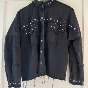 Svart jeansjacka med nitar - Säljer en cool svart jeansjacka med nitar och fransiga detaljer. Jackan har en avslappnad passform och knappar framtill. Perfekt till när det år lite hostigt kallt väder. Storlek xs men sitter som en S