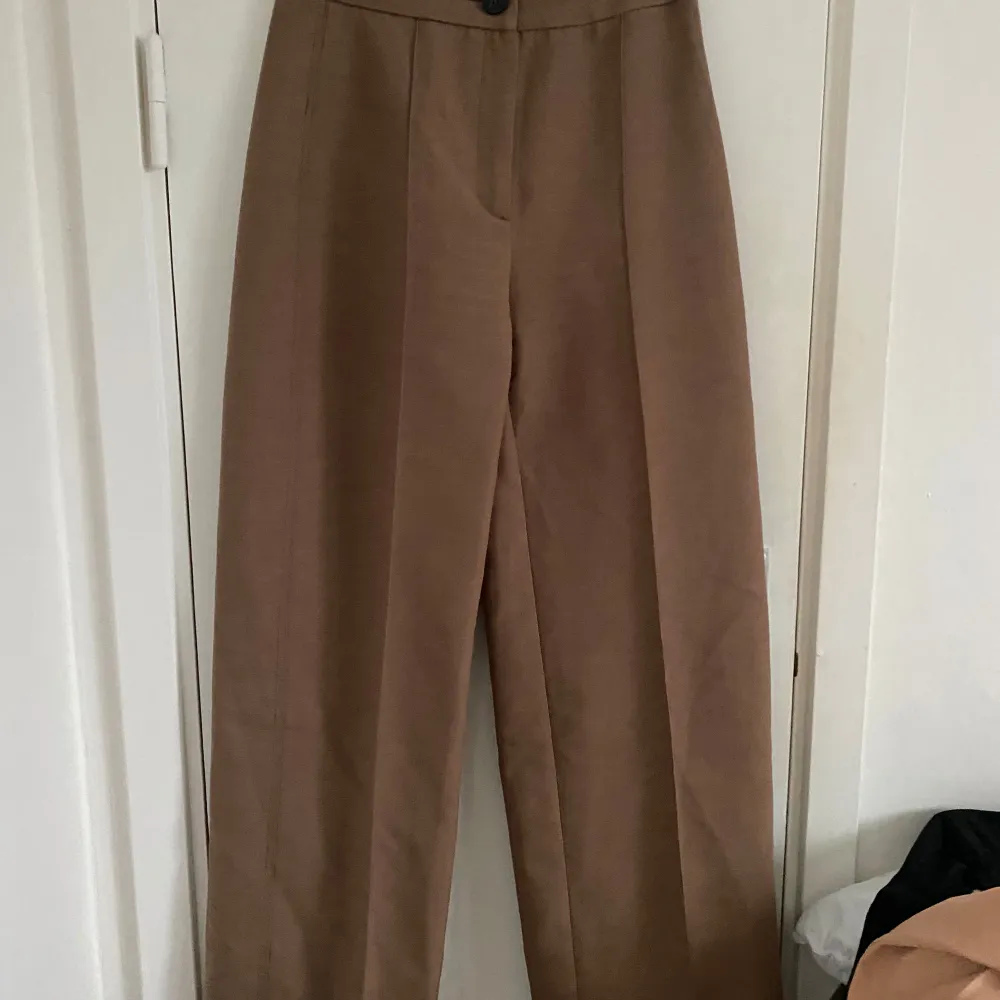 Snygga beige kostymbyxor från Bershka i storlek EUR 36. De har en hög midja och en klassisk rak passform. Perfekta för både kontoret och mer formella tillfällen. Byxorna har en knapp och dragkedja framtill för enkel stängning.. Jeans & Byxor.