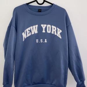 Blå sweatshirt med New York-tryck - Säljer en snygg blå sweatshirt med texten 'New York U.S.A' i vitt på framsidan. Tröjan har en avslappnad passform och är perfekt för både vardag och chill. Den är i bra skick och superbekväm att ha på sig. Perfekt för höst och vår!