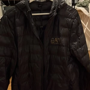 Svart/gold  dunjacka från Emporio Armani -  Säljer en snygg svart dunjacka från Emporio Armani. Jackan har en quiltad design med dragkedja framtill o huva och en påse i jackan du kan läggga ner den i vid eventuella resor osv  och EA7-logga på bröstet. Perfekt för kyliga dagar och ger en stilren look. Passar både till vardags och lite finare tillfällen. äkta såklart NYPRIS 1700 