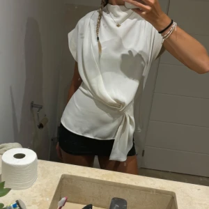 Vit blus med knytning - Säljer en stilren vit blus med knytning i midjan. Den har en elegant design med korta ärmar och en hög hals. Perfekt för både vardag och festliga tillfällen. Passar bra till både jeans och kjol!