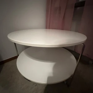 Vit rund sidobord med två hyllplan - Säljer ett stilrent vitt sidobord med två runda hyllplan. Bordet har en modern design med metallben som ger en stabil känsla. Perfekt för vardagsrummet eller som nattduksbord. Bordet är i gott skick och passar in i de flesta inredningsstilar. Perfekt för att ställa böcker, växter eller dekorativa föremål på. 