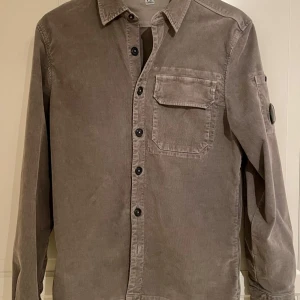 C.P. Company Corduroy Lens Overshirt - Ursnygg skjorta i smalspårigt manchestertyg från CP Company. Storlek Small, sitter bra på mig som är 176cm.  Väldigt fint skick, inget att anmärka på. Tags, etiketter och extra knappar medföljer. Skjortan är som denna (https://www.cpcompany.com/en-se/green-corduroy-lens-buttoned-shirt/17CMSH160A006410O674.html?dwvar_17CMSH160A006410O674_color=674&dwvar_17CMSH160A006410O674_size=L&quantity=1) men med dold knäppning på bröstfickan (snyggare!) och ljusgrå. 