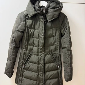 Olivgrön dunjacka från Rockandblue - Säljer en olivgrön dunjacka från Rockandblue i storlek 34. Jackan har en lång och figursydd design med dragkedja och knappar framtill. Den är perfekt för kalla vinterdagar med sin varma vaddering och huva. Två praktiska fickor med dragkedja på sidorna. Superfin och i mycket bra skick! Päls finns. 