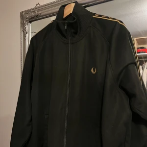Svart jacka från Fred Perry - Säljer en snygg svart jacka från Fred Perry i mycket bra skick. Den har en dragkedja framtill och coola detaljer med Fred Perry-loggan längs ärmarna. Perfekt för höst och vår, och den har en normal passform. Jackan är långärmad och gjord i ett bekvämt material.