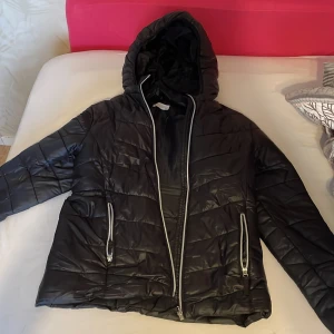 Svart quiltad pufferjacka - Säljer en svart quiltad pufferjacka med huva. Jackan har dragkedja framtill och två fickor med dragkedjor. Perfekt för kyliga dagar och håller dig varm och bekväm. Passar både till vardags och lite mer avslappnade tillfällen.