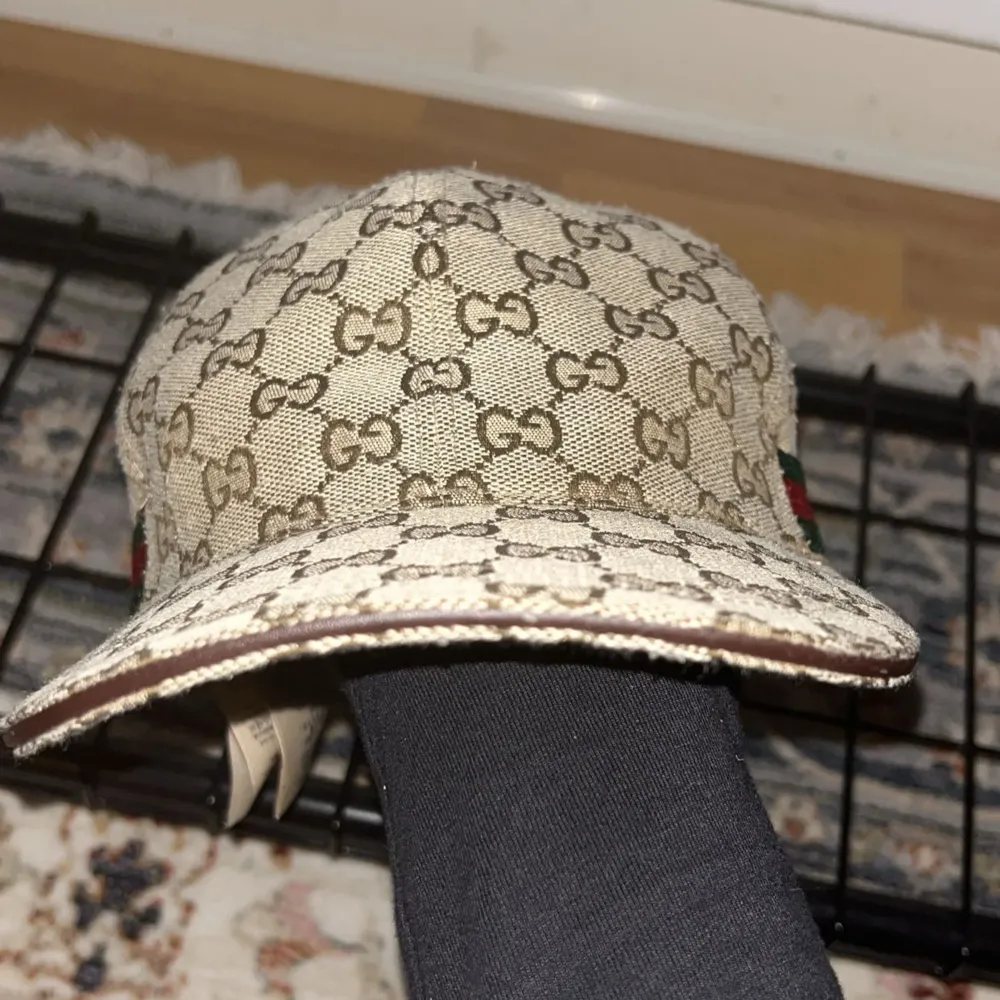 Säljer en snygg beige keps från Gucci med deras ikoniska GG-mönster. Kepsen har en klassisk design med en röd och grön randdetalj på sidan. Perfekt för att ge din outfit en lyxig touch. Tillverkad i Italien och i mycket bra skick.. Accessoarer.