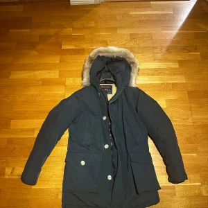Woolrich arctic Mörkblå - Säljer en snygg mörkblå parkajacka med beige quiltat foder och pälsdetalj på huvan. Jackan har praktiska fickor och knappar framtill. Perfekt för kalla vinterdagar! 🧥❄️