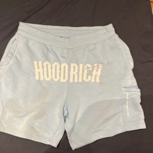 Ljusblå shorts från Hoodrich - Säljer ett par ljusblå shorts från Hoodrich i storlek L. De har en cool design med tryck på både fram- och baksidan. Shortsen har en praktisk ficka med dragkedja på sidan. Perfekta för en avslappnad stil i sommar!