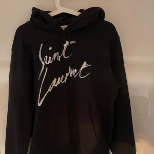 Svart hoodie från Saint Laurent - Säljer en snygg svart hoodie från Saint Laurent med deras ikoniska logga i vitt på framsidan. Den är i mycket bra skick och perfekt för både höst och vinter. Hoodien har en klassisk passform med långa ärmar och en bekväm huva. Perfekt för en stilren och avslappnad look!