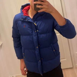 Blå pufferjacka från Polo Ralph Lauren - Säljer en snygg blå pufferjacka från Polo Ralph Lauren i storlek L i ungdomsstorlek. Jackan har en röd insida och är perfekt för kalla dagar. Den har både dragkedja och knappar framtill för extra skydd mot kylan. Perfekt för vintern! ❄️